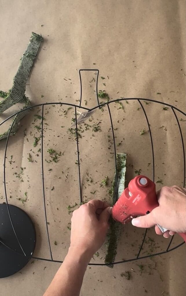 A person uses a hot glue gun to attach a strip of green fabric to a metal pumpkin-shaped frame in this Dollar Tree pumpkin wreath DIY. The frame and fabric strips are placed on a brown surface, with some green fabric trimmings scattered around.