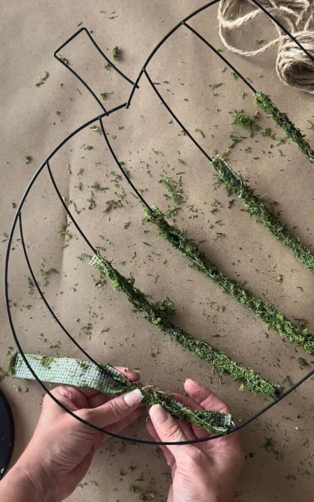 Hands wrapping green moss around sections of a wire pumpkin frame, with pieces of moss and a spool of twine visible on the table. The Dollar Tree pumpkin wreath DIY project appears to be a craft in progress.