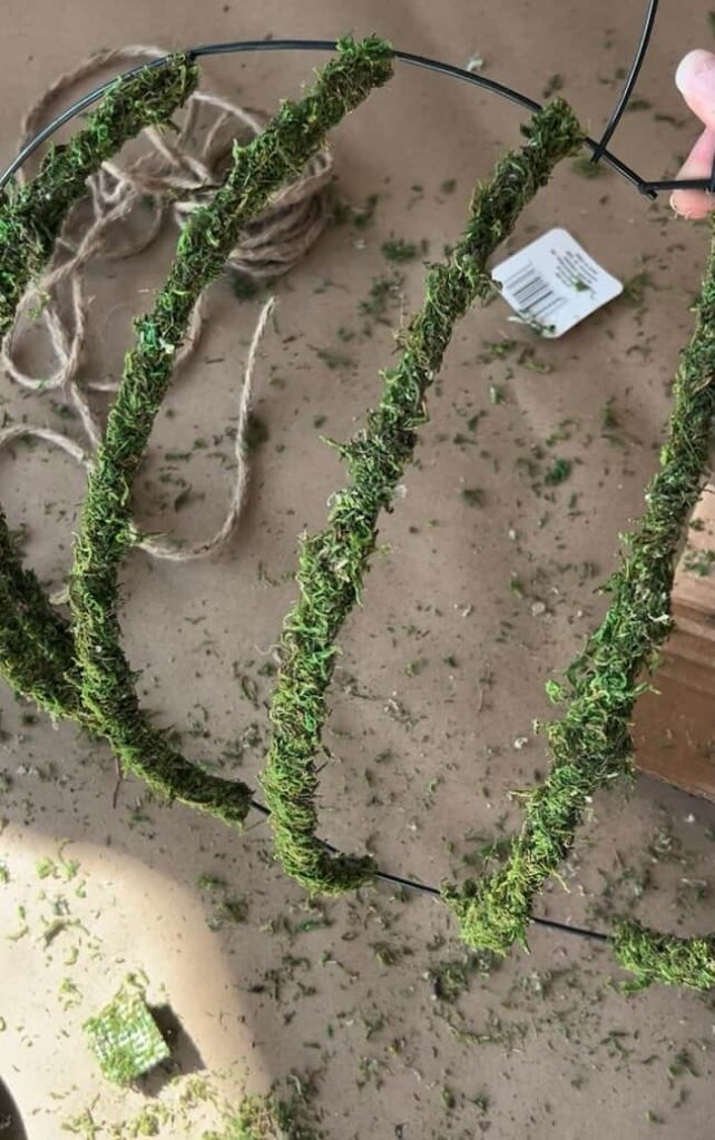 A hand holds a wire Dollar Tree pumpkin wreath frame partially covered in green moss on a messy surface. Loose moss and biodegradable materials are scattered around, including a piece of twine and small plastic packaging. The crafting process appears to be in progress.