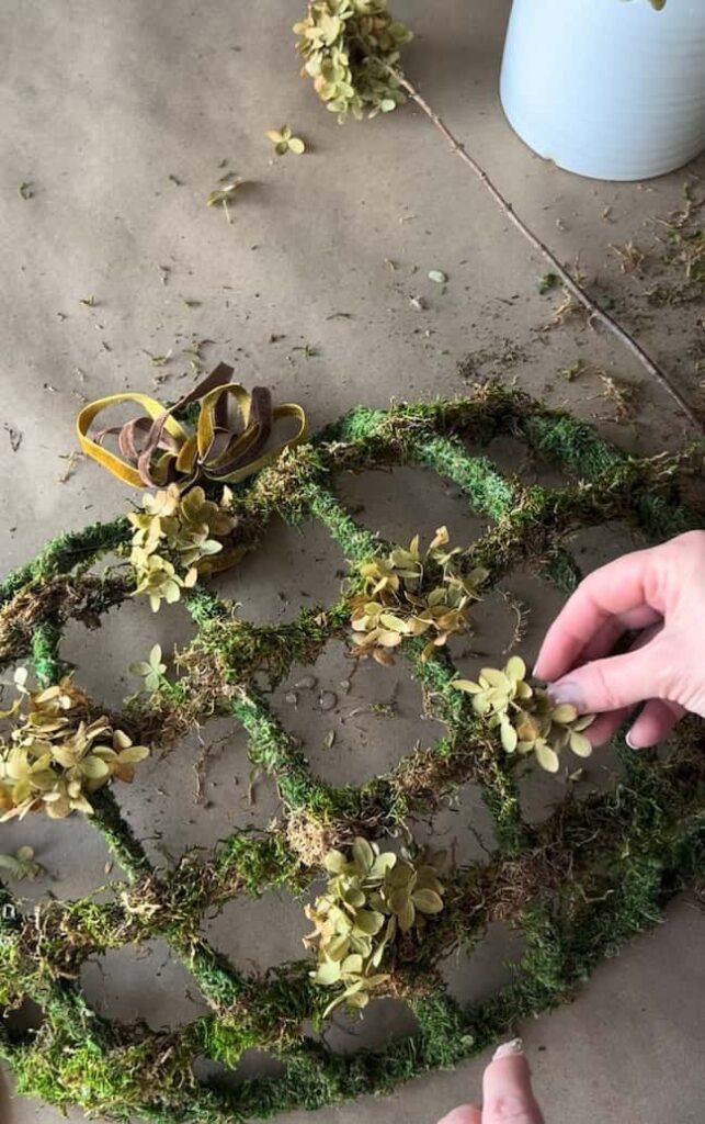 Two hands work on a circular craft project covered in green moss and adorned with dried flowers. A pair of scissors and a bunch of ribbon are nearby on a brown surface, reminiscent of a Dollar Tree pumpkin wreath DIY. A white container is partially visible in the top right corner.