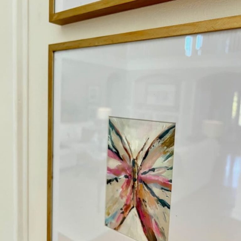 A framed abstract painting with vibrant splashes of pink, black, and gold lines hangs on a white wall. The painting appears to be part of a gallery display, with another frame partially visible above it. Reflections in the glass show a blurred room in the background, suggesting a recent Picture Frame Makeover.