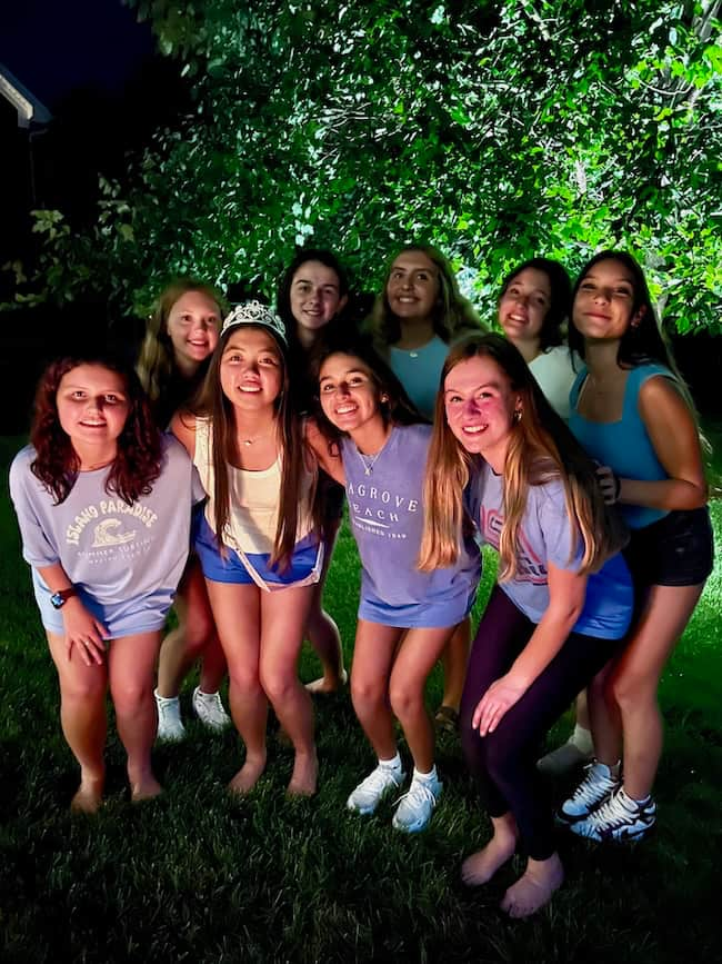 A group of nine young women are standing closely together outdoors at night, smiling at the camera. They are dressed in casual summer clothing, and one woman in the front is wearing a tiara. The background features greenery, illuminated by lights. It's a Seven on Saturday to remember!