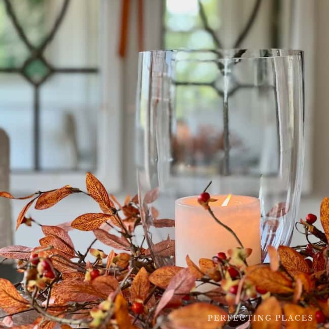 A lit white candle inside a tall glass vase is surrounded by orange and red autumn leaves and berries. The arrangement, perfect for fall decoration, is set on a table with a blurred, white-framed window in the background. Mark your calendars for the event date 9/21/24 – Seven on Saturday awaits!