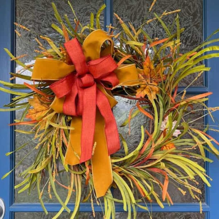 A festive autumn wreath made of green, yellow, and orange foliage with a large, vibrant orange bow is hanging on a blue door with a textured glass insert. This easy fall wreath DIY adds the perfect seasonal touch to your home.