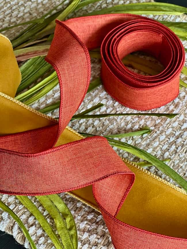 A roll of red ribbon is uncoiled next to another partially uncoiled roll of yellow fabric ribbon. They are laid out on a woven surface, with long green leaves scattered around them—perfect for an easy fall wreath DIY project.