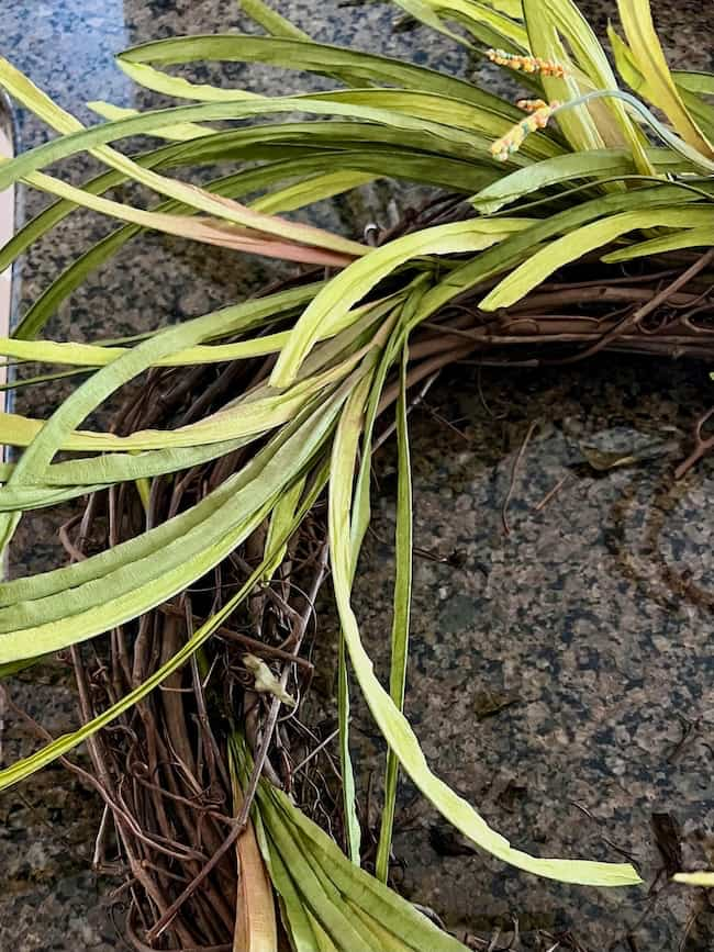 A close-up of a decorative wreath made from intertwined twigs and adorned with green artificial leaves and small yellow flowers. The easy fall wreath DIY project is laid on a textured granite surface.