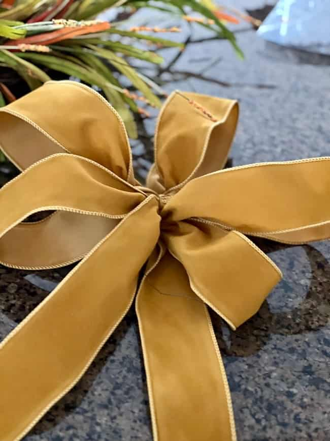 A close-up of a neatly tied yellow ribbon bow with long tails, placed on a granite surface, serves as the perfect centerpiece for an easy fall wreath DIY. The background includes some out-of-focus green foliage.