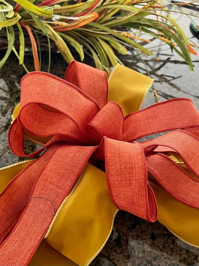 A decorative bow made of orange and yellow ribbon on a reflective surface, with green foliage and some orange accents in the background. The bow has multiple loops and adds a festive touch to the easy fall wreath DIY scene.