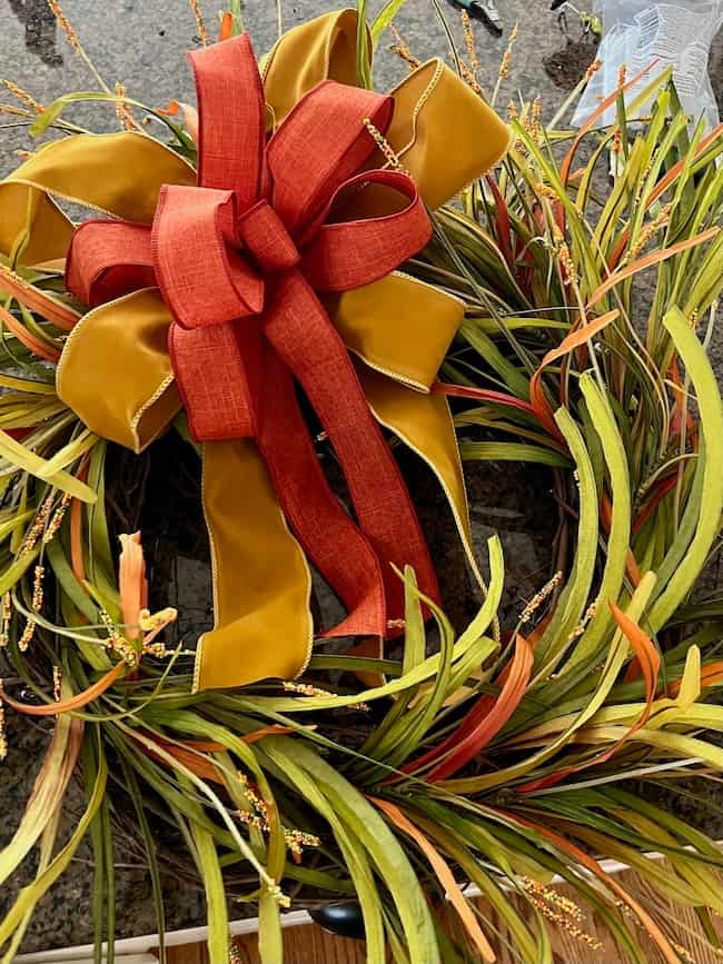 A decorative wreath made of green and orange foliage, adorned with a large bow featuring red and yellow ribbons, placed on a gray surface. The wreath, perfect for an easy fall DIY project, has a natural, autumnal appearance.