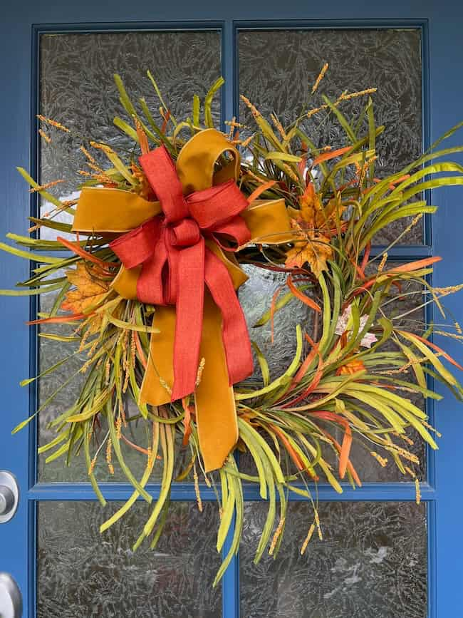 A decorative autumn wreath on a frosted glass front door. The easy fall wreath DIY project is adorned with green and orange foliage, yellow accents, and features a large orange and yellow bow at the top. The door is painted blue.