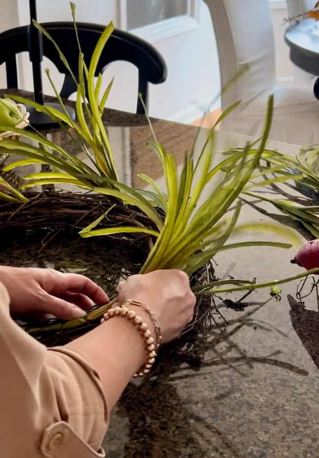 A person is creating an easy fall wreath DIY by arranging light green foliage into a brown wreath frame on a brown speckled countertop. The person's hands, adorned with a beaded bracelet, are visible. A black chair sits in the background.