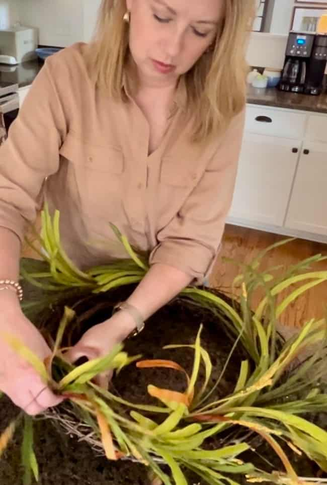 A person with blond hair in a beige shirt is making an easy fall wreath with greenery at a kitchen counter. The counter has a dark granite top with kitchen appliances and cabinets visible in the background.