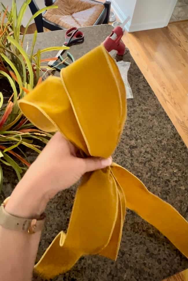 A person is holding a yellowish-orange ribbon with one hand on a textured dark countertop while crafting an easy fall wreath DIY. A hot glue gun and a potted plant rest nearby on the counter, with a wooden floor visible in the background.