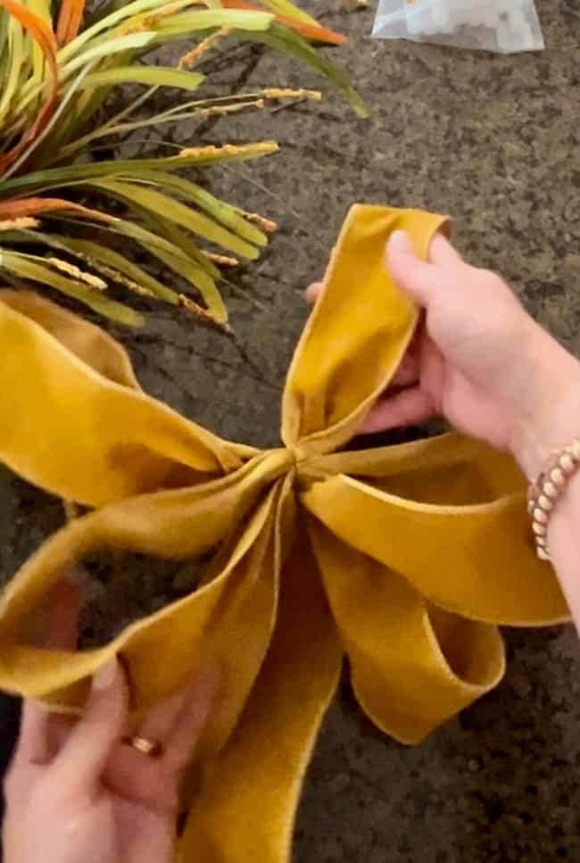 A pair of hands is tying a large, yellow fabric ribbon into a bow on a dark textured surface, perfect for an easy fall wreath DIY. An arrangement of green and yellowish decorative foliage is partially visible in the background. The person is wearing a bracelet on one wrist.