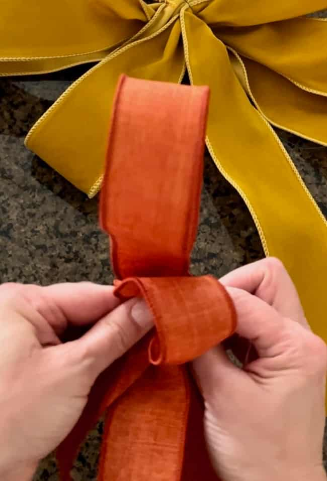 Close-up of hands crafting a bow with an orange ribbon. A completed bow made from yellow ribbon is placed on a dark, speckled surface in the background—perfect accents for an easy fall wreath DIY project.