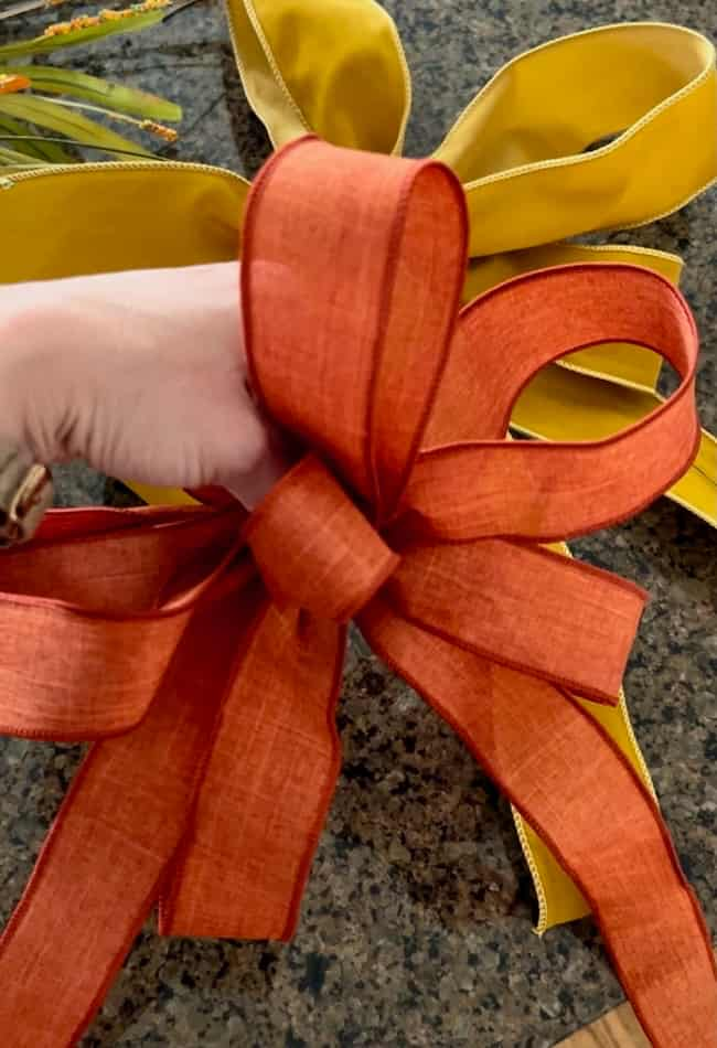 A hand is holding an orange bow with multiple loops and tails. In the background, a yellow bow of similar style is visible, lying on a dark speckled surface. The scene appears to be related to craft or gift wrapping, perfect for an easy fall wreath DIY project.