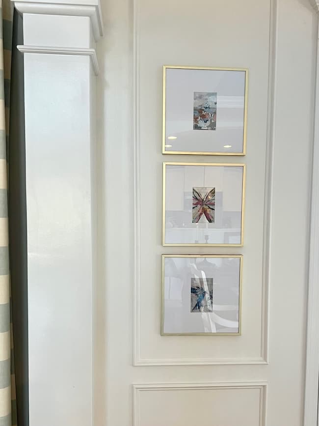 A white wall features three framed abstract art pieces arranged vertically. Each artwork is in a gold frame and shows a colorful, abstract pattern. The wall has decorative molding, and on the left side, a doorway with a striped curtain is partially visible. The frames underwent a Rub 'n Buff makeover for added elegance.