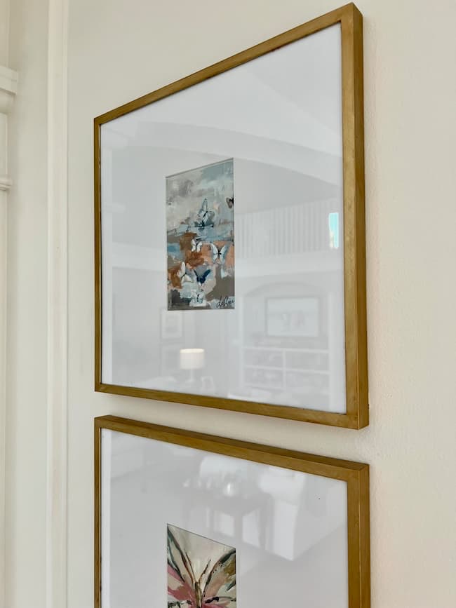 Two wooden-framed abstract paintings hang on a light-colored wall. The upper painting features a mix of blue and brown tones, while the lower one showcases a blend of pink, green, and white colors. Both frames, enhanced with a Rub N Buff Picture Frame Makeover, are aligned vertically on the wall.