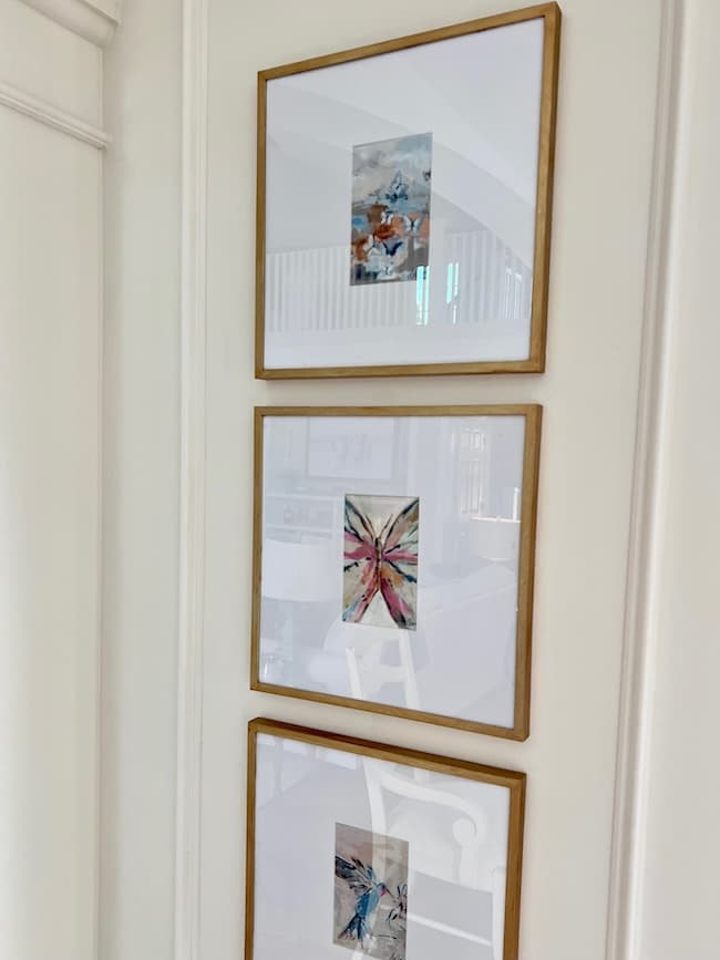 Three framed abstract art pieces are arranged vertically on a light-colored wall. Each frame features a brassy to classy, thin gold border and displays a unique, colorful abstract painting against a white matte background. The setting appears to be an interior space with bright lighting.