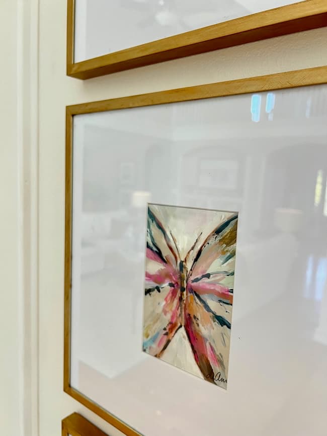 A framed abstract painting of a butterfly in vibrant colors, including pink, black, blue, and beige. The golden frame has the elegance of a Rub N Buff Picture Frame Makeover. Mounted on a wall in a well-lit room with blurred furniture in the background, the signature "Ana" graces the bottom right.