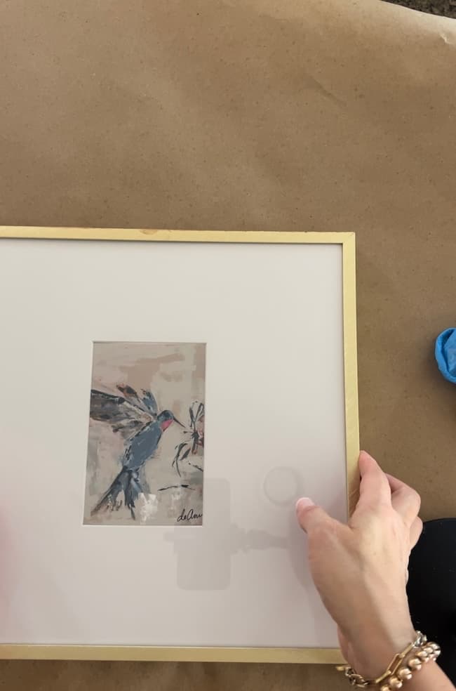 A person is holding a framed painting of a bird with their hands. The painting has a white background with a gold frame, accentuated by Rub 'n Buff. The person's wrists are adorned with bracelets, and the picture frame makeover beautifully complements the brown surface it rests upon.