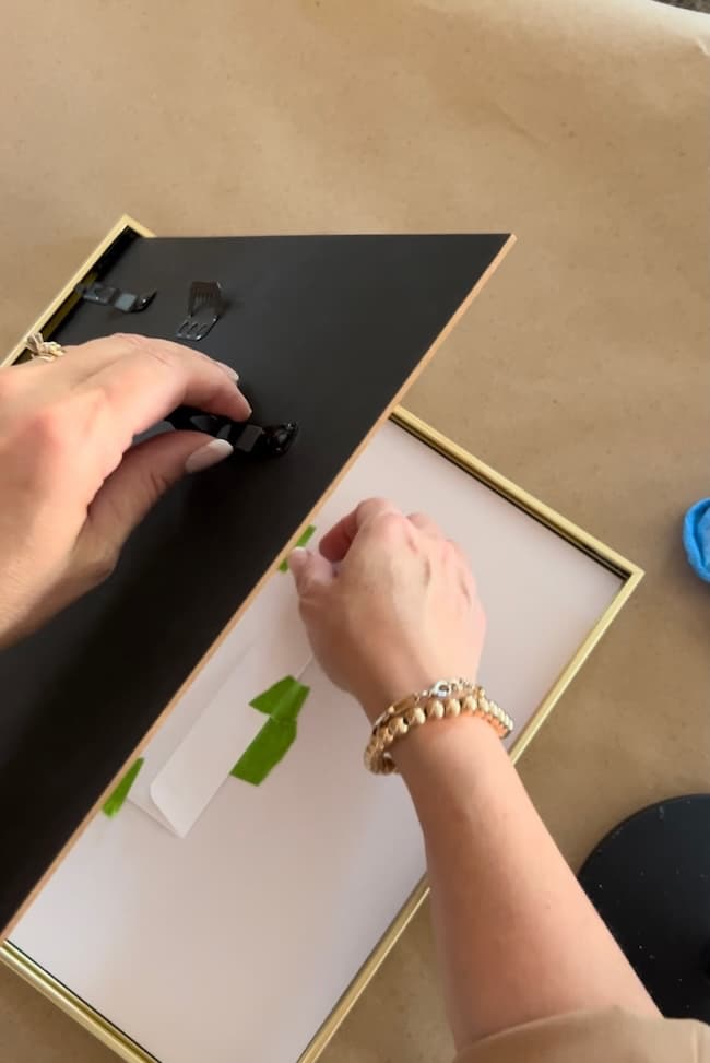 A person is seen from their wrist down, using both hands to remove the backing of a picture frame for a picture frame makeover. The frame lies on a table covered in brown paper. The individual, wearing a beige long-sleeve shirt and gold bracelet, appears ready to transform the piece from brassy to classy.