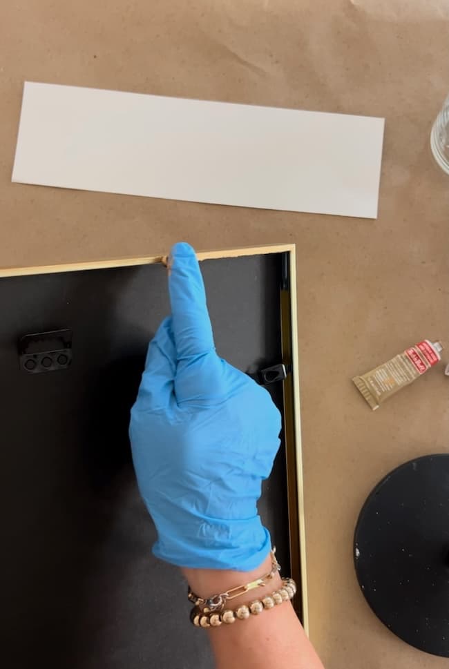 A person wearing a blue glove is holding the corner of a picture frame with a gold edge, possibly prepping for a picture frame makeover. A rectangular white piece of paper is placed on the brown surface beside the frame. The person is also wearing a bracelet with white beads, implying DIY creativity in progress.