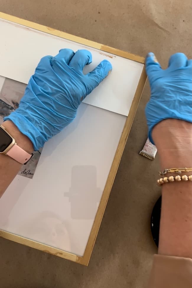 Person wearing blue gloves and a wristwatch is adjusting a picture in a golden frame, likely part of a Rub N Buff Picture Frame Makeover. A tube of adhesive lies nearby on the tan surface. The frame holds a photograph, and part of another wrist, with a bracelet, is visible.