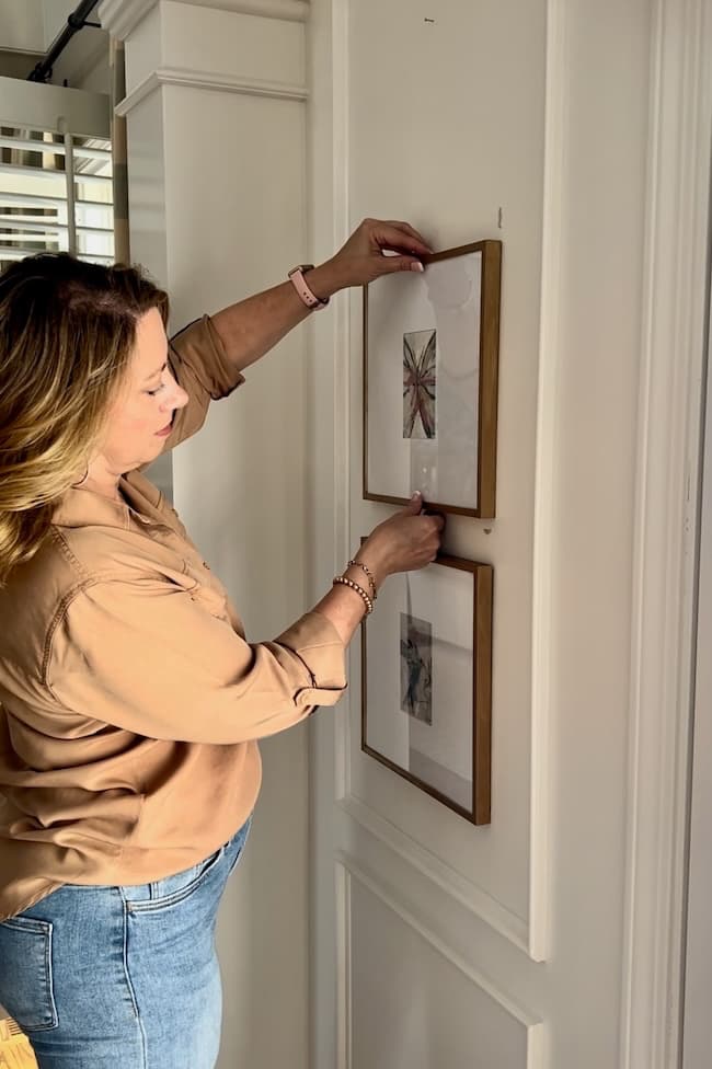 A woman with light brown hair is hanging two framed pictures on a white wall. She is wearing a beige blouse and blue jeans, carefully adjusting the top frame for a Brassy to Classy Picture Frame Makeover.