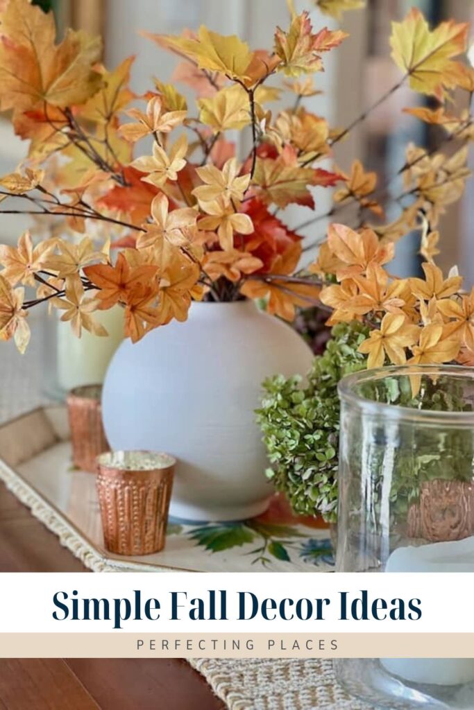 A white vase filled with yellow and orange autumn leaves is placed on a tray. Next to the vase are small copper candle holders and a green plant. The text at the bottom reads "Perfecting Places" with a subheading of "Simple Fall Decor Ideas.