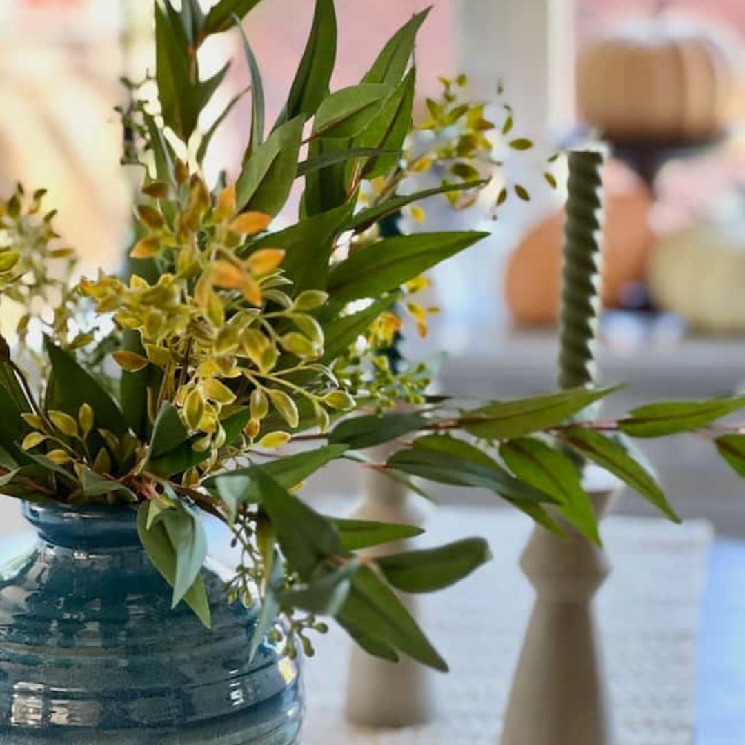 A ceramic vase filled with green and yellow foliage is placed on a table. Next to it, there's a green twisted candle in a holder. The background is softly blurred with warm tones, and there are pumpkins visible, suggesting cozy, simple fall decor ideas.