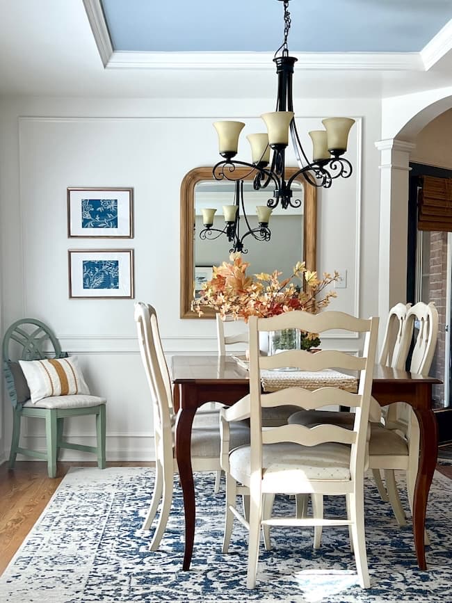 A dining room with a wooden table surrounded by white chairs. A chandelier with multiple lights hangs above the table. A floral centerpiece sits on the table. The walls feature a large mirror and two framed artworks, perfect for Simple Decor Ideas. A green chair and cushion sit cozily in the corner.