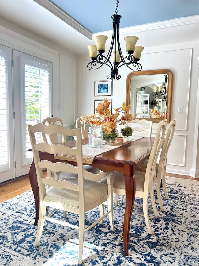 A cozy fall home tour showcases a dining room with a wooden table and six white chairs. The table is adorned with fall-themed decorations, including orange and yellow foliage arrangements. A large mirror hangs on the wall, reflecting light from the nearby window. Above, a chandelier with five lights casts a warm autumn glow.