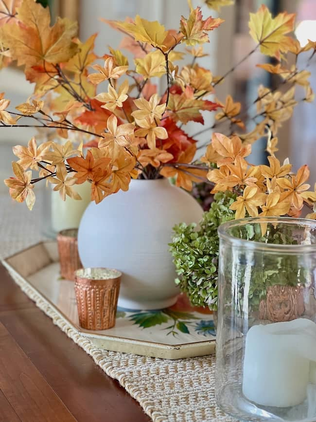 A table adorned with a decorative autumn centerpiece featuring a white vase filled with orange and yellow fall leaves. Adding to the simple decor, the arrangement is complemented by small copper candle holders and a large glass candle holder, all resting on a woven table runner for that cozy fall home touch.