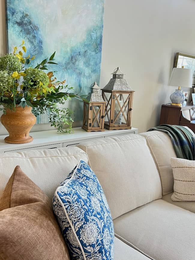 A cozy living room features a cream-colored sofa adorned with brown, blue, and white patterned pillows. Behind the sofa, a console table displays a potted floral arrangement, two lanterns, and a table lamp. For a Cozy Fall Home Tour feel, a large abstract blue and green painting hangs on the wall.