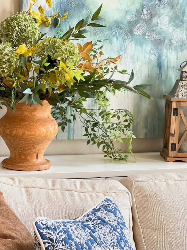 A cozy fall home setup featuring a white couch adorned with a blue-patterned pillow. Behind the couch sits a large terracotta vase filled with an arrangement of green and yellow foliage. A lantern decorates the surface beside the vase, and a textured blue painting hangs on the wall, perfect for simple decor ideas.