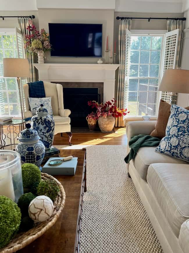 A cozy living room with a fireplace, TV mounted above, and large windows. The room features a beige sofa with blue-patterned cushions, decorative vases, and a woven basket with green moss balls on the coffee table. For your Cozy Fall Home Tour, tall lamps flank the fireplace for that warm autumn refresh.