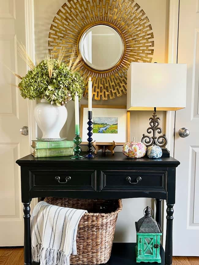 A black console table holds a white vase with green hydrangeas, candles, a lamp, and decorative items. Below, a woven basket with a striped blanket and a green lantern sits. Above, a round sunburst mirror hangs on the wall. For cozy fall home vibes, incorporate these simple decor ideas into your space.