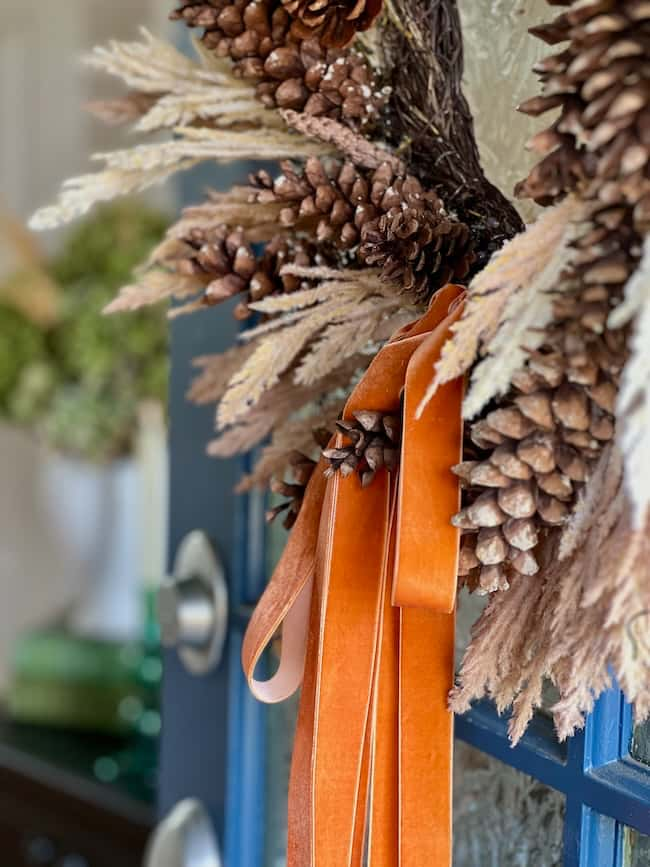 Close-up of a fall-themed wreath on a door. The wreath includes pinecones, dried foliage, and long orange ribbons, creating a festive autumn look. Simple decor ideas like this add to the cozy fall home ambiance. The door's handle is visible, hinting at an inviting entrance.