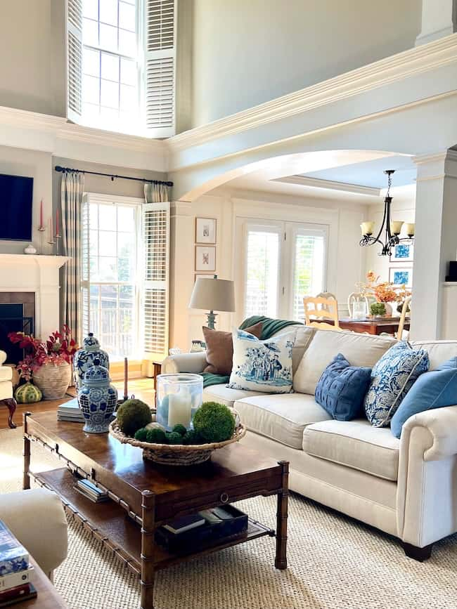 A spacious living room with high ceilings and large windows allowing natural light to flow in, perfect for a Cozy Fall Home. The room features a white sofa adorned with blue-patterned pillows, a wooden coffee table with decorative items, and a dining area in the background. The decor is elegant and inviting.