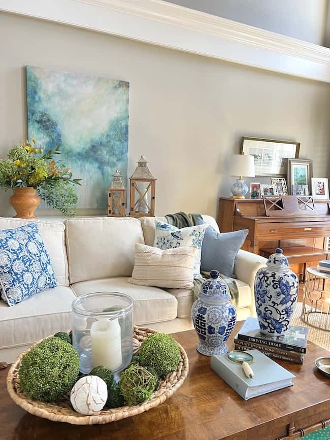 A cozy living room featuring a beige couch with blue and white patterned pillows. A wooden coffee table holds decorative greenery and candles, while ginger jars and books sit nearby. In the background, there's a console table with family photos and a piano adorned with art for a warm autumn refresh.