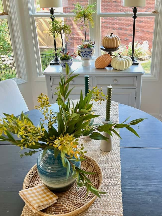 A cozy fall home dining area features a table with a round woven placemat, a blue vase with yellow flowers, two green candles in holders, and a yellow checked napkin. In the background, there is a sideboard by a window, decorated with ornamental gourds and indoor plants for that warm autumn refresh.