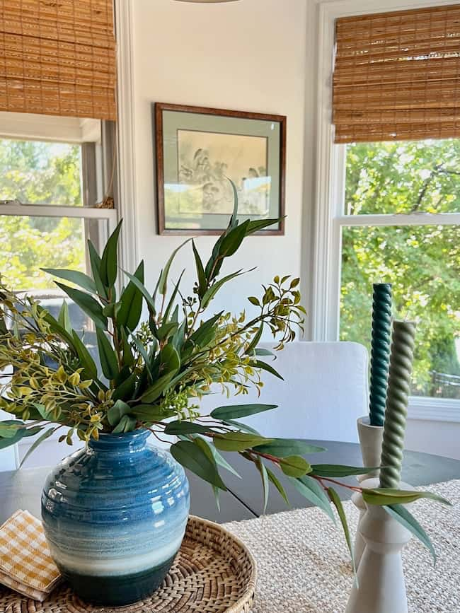 A cozy dining area with large windows allowing natural light. A blue ceramic vase with green leaves and yellow flowers sits on a woven placemat on the table, exemplifying simple decor ideas. Near it, two twisted candles, one green and one blue, stand in white holders. A framed artwork hangs on the wall.