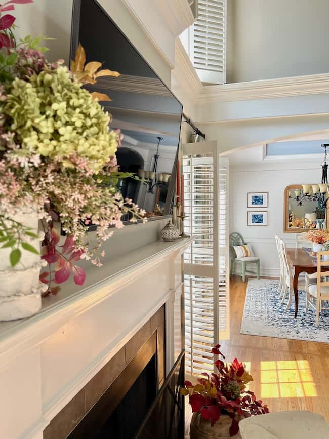 A bright living room features elegant white woodwork, a TV above the fireplace, and floral arrangements on the mantel and floor. Sunlight streams through partially opened plantation shutters, leading into a dining area with a large mirror and chandelier—perfect for your cozy fall home aesthetic.