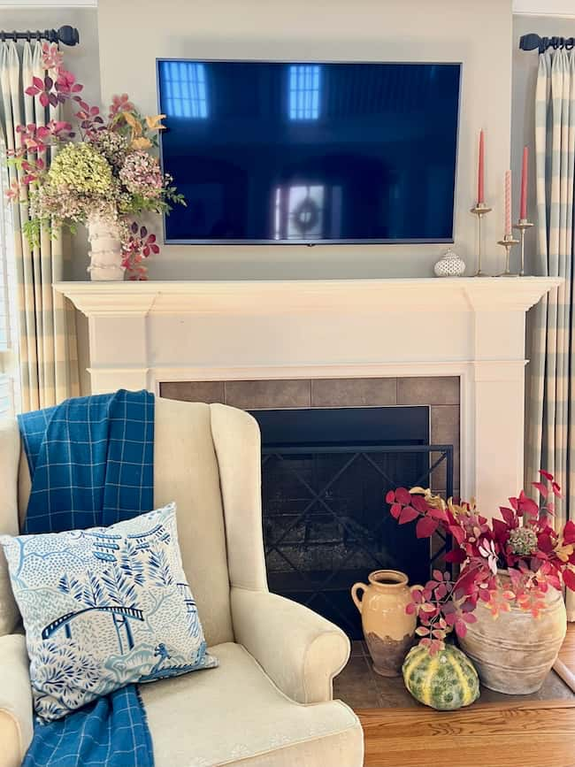 A cozy living room features a fireplace with a TV mounted above it. Floral arrangements and candles decorate the mantel. Below, two vases with flowers and pumpkins, perfect for a Warm Autumn Refresh, sit beside the hearth. An armchair with a blue throw and patterned pillow is positioned nearby.