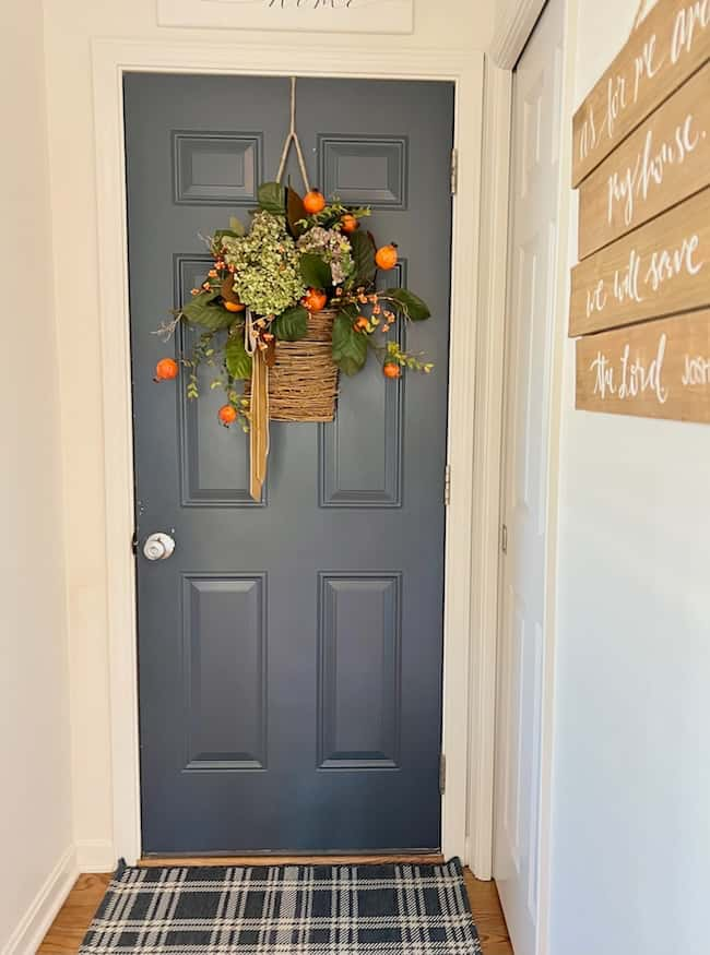 A blue door adorned with a basket wreath filled with greenery, white flowers, and small orange fruits. The door is set in a narrow hallway with white walls, a plaid rug on the floor, and wooden wall decor featuring inspirational quotes on the right side—a perfect touch for Cozy Fall Home vibes.