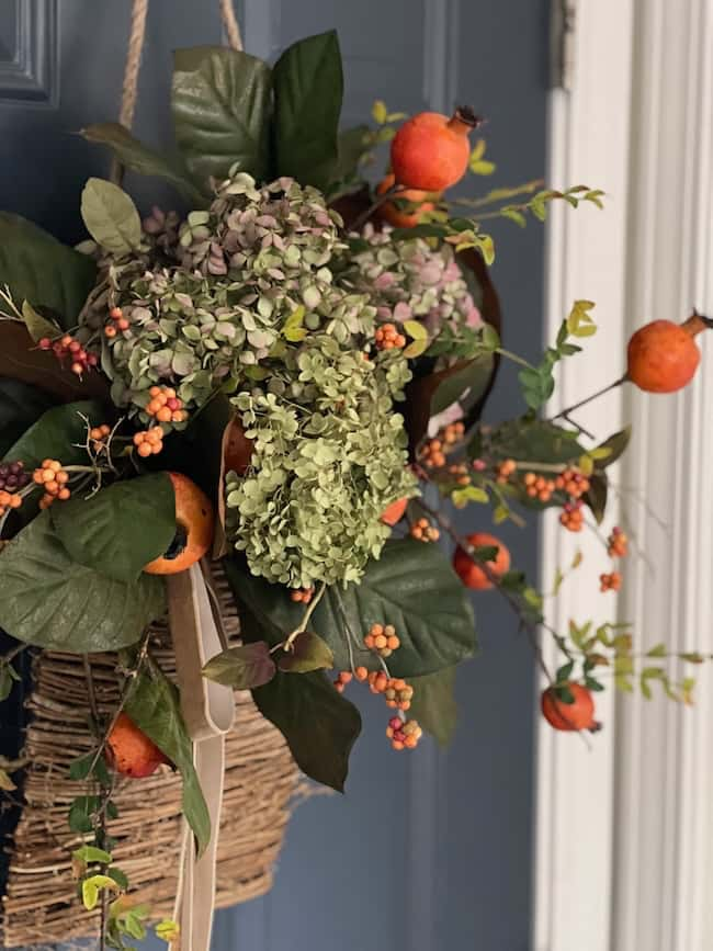 A decorative hanging basket is filled with a lush arrangement of greenery, hydrangeas, orange berries, and small orange fruits, possibly pomegranates. This Cozy Fall Home accent adds charm against the dark blue door.