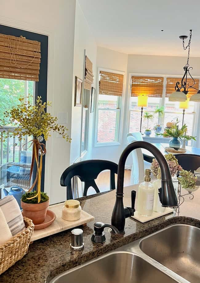 A bright kitchen with a granite countertop featuring a black faucet, soap dispenser, and potted plant provides Simple Decor Ideas. A wicker basket sits on the counter. The kitchen opens into a dining area with a window, wicker blinds, and a table with a chandelier overhead.