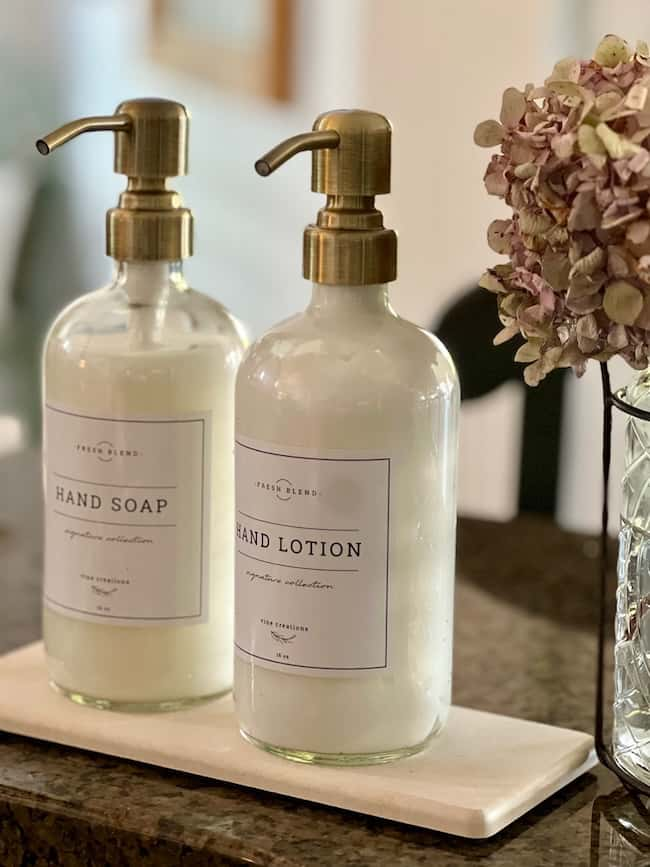 Glass bottles with brass pumps filled with hand soap and hand lotion sit on a tray on the bathroom counter. The bottles are labeled "Hand Soap" and "Hand Lotion." A decorative vase with dried pink hydrangeas adds to the cozy fall home vibe.