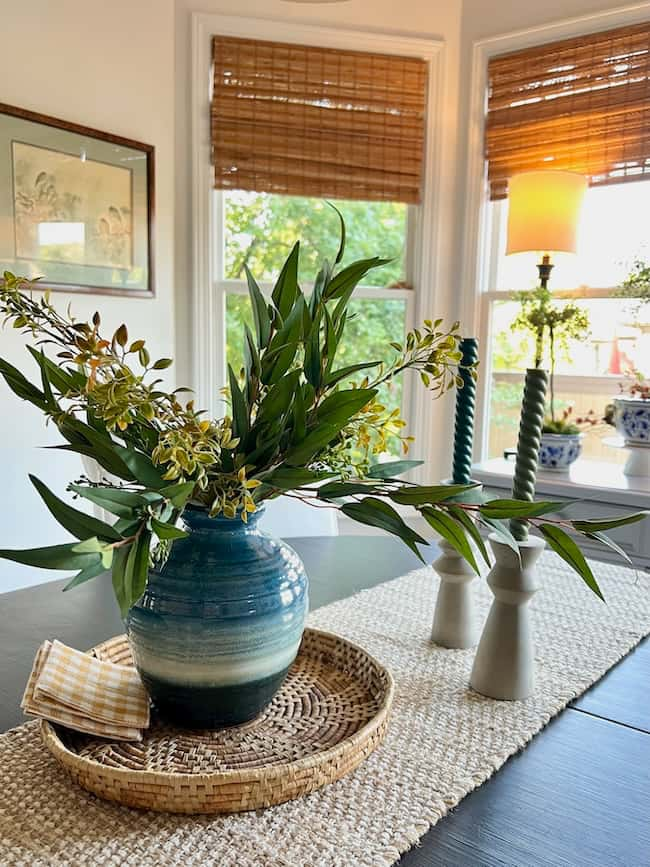 A blue-green gradient vase with leafy branches and yellow flowers sits on a woven tray on a table runner. Behind are bamboo blinds on windows, a lit lamp, and candlesticks with green spiral candles, creating a cozy fall home ambiance—a perfect warm autumn refresh for simple decor ideas.